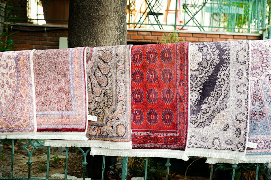 A row of colorful Persian-style rugs laid out outdoors on a metal railing for drying or airing, each showcasing intricate patterns in shades of red, beige, black, and gray. The rugs appear clean, with