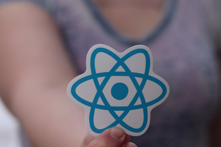A person is holding a small cut-out paper sticker featuring a stylized blue atomic symbol with overlapping elliptical orbits and a central circle, against a blurred background of a person wearing a pu